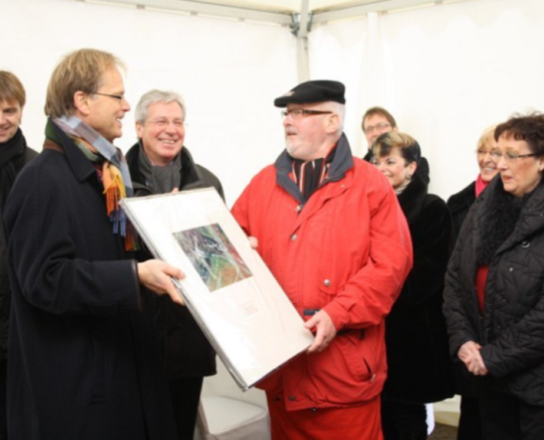 Ein Mann überreicht einem anderen Mann einen Bilderrahmen. Im Hintergrund stehen weitere Menschen. | © Wilfried Moritz/Magistrat der Stadt Bremerhaven