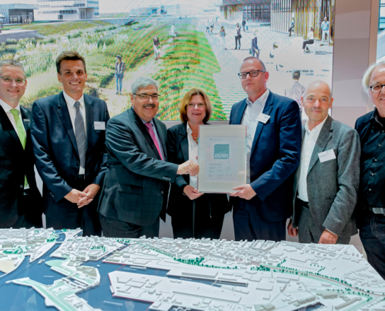 Sechs Männer und eine Frau lächeln in die Kamera, zwei Männer halten ein DIN A4 großen Zettel in die Kamera. Im vordergrund ist ein Modell einer Stadt zu sehen. | © BIS/Frank Pusch