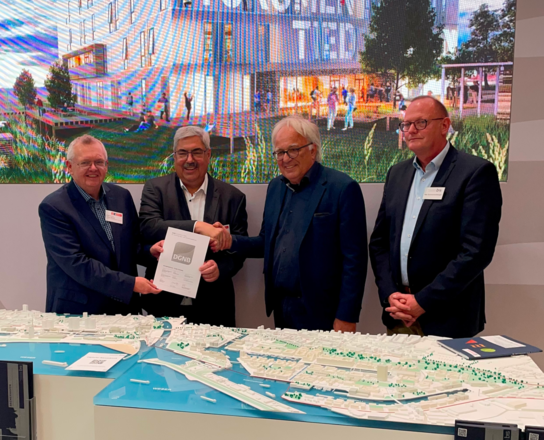 Vier Männer lächeln in die Kamera vor einem Stadtmodell. Zwei der Männer halten einen DinA4 großen Zettel in die Kamera. Im Hintergrund ist eine Leinwand. | © BIS Bremerhaven